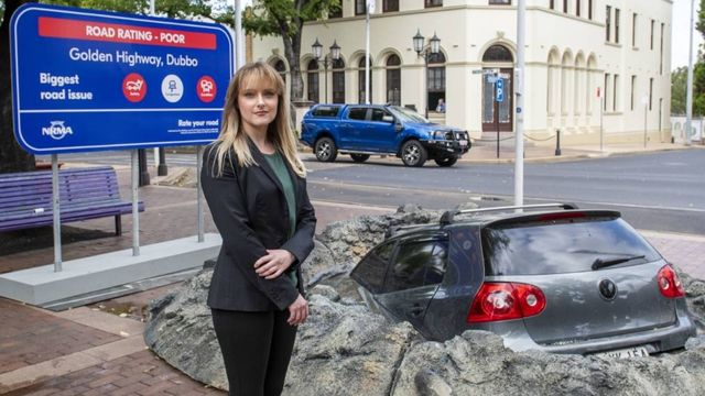 woman wearing business attire standing next to a grey vehicle stuck in a giant pothole