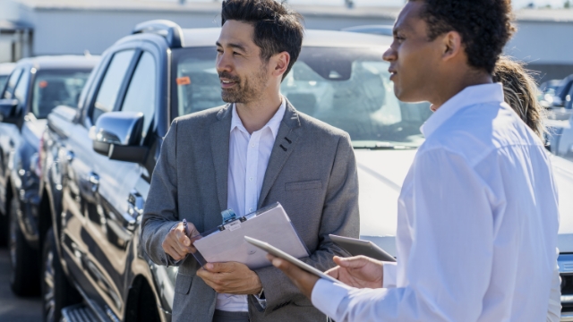 two men in business clothing standing in front of a line of cars looking sideways