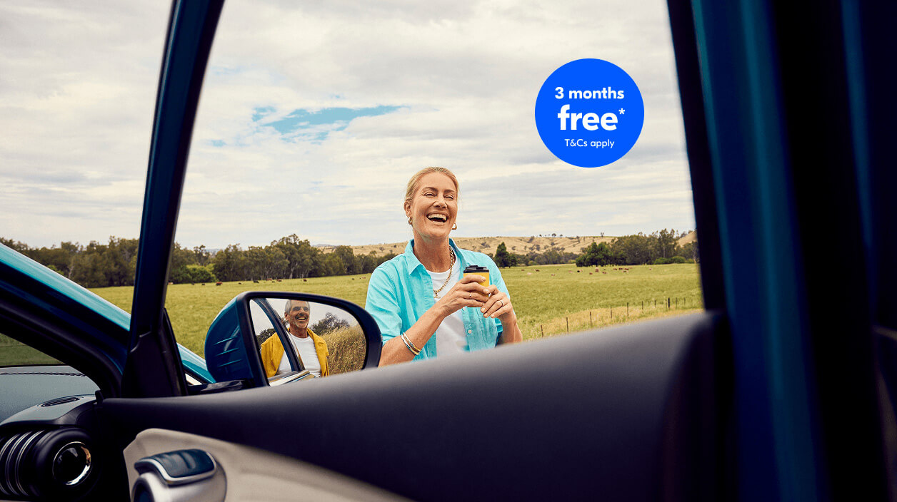 A smiling woman standing outside her car in a rural field, holding a drink, pictured from inside the vehicle