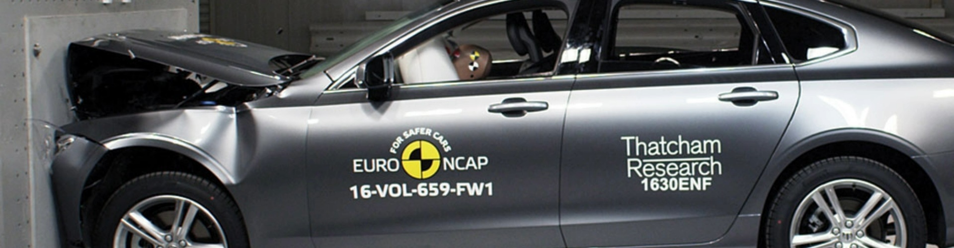 Side view of a grey car in a head-on collision with a wall during a crash test.