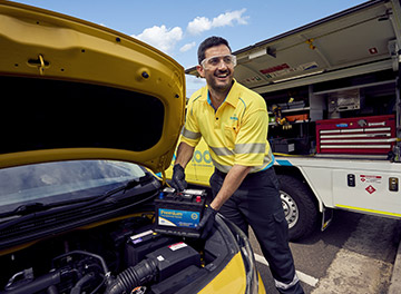 A Roadside assistance NRMA staff member in uniform holding a car battery in his hands smiling while standing next to a vehicle with an open bonnet