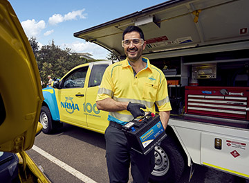 A Roadside assistance NRMA staff member in uniform smiling while holding a large battery in his hands, showcasing its size and importance.
