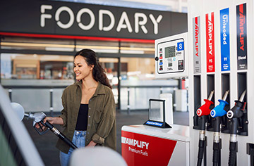 A woman at an Ampol fuel station is filling her car's tank, holding the fuel nozzle and standing beside her vehicle.