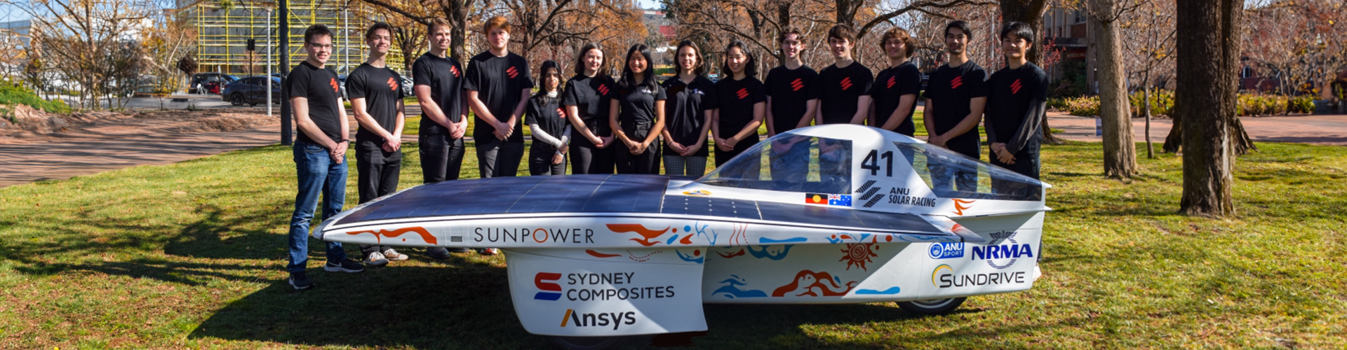 A group of fourteen university students dressed in black t-shirts, stand behind a small, car-like vehicle with solar panels along the hood, and the word Sunpower along the trim. A group of fourteen university students dressed in black t-shirts, stand behind a small, car-like vehicle with solar panels along the hood, and the word Sunpower along the trim.