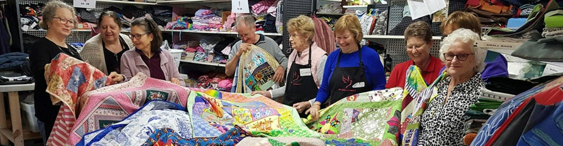 local-hero-derryk-and-jackie-1920x500 A group of older women and one man gathered around a table full of handmade quilts in an op shop.