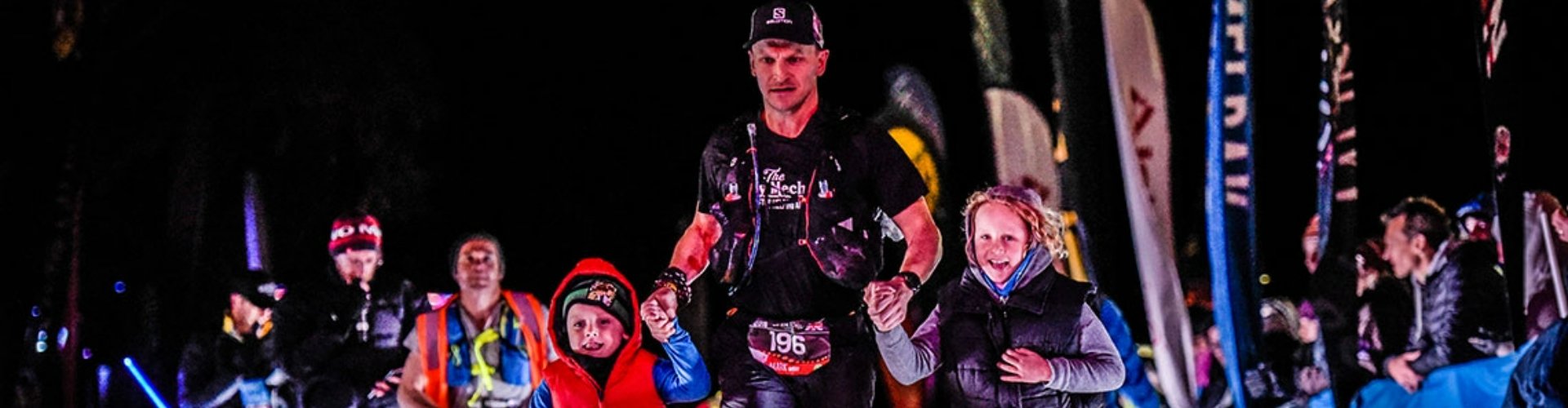 local-hero-mark-avery-1920x500 A man finishes a marathon at night, with his two children by his side as he runs past crowds.
