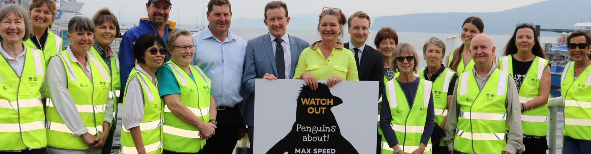 manly-penguins-npws-nrma-1920x500 A man in a suit and a woman holding a sign saying, Watch out penguins about, surrounded by volunteers in yellow high vis jackets.
