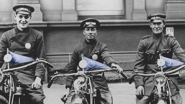 black and white photo from the early 1920s showing a row of four young NRMA patrolmen on their motorbikes that have blue triangular NRMA flags on the handlebars