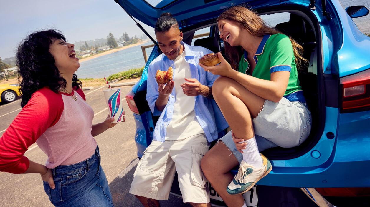 Three people gather by the open trunk of a blue hatchback near a beach, holding pies and a drink
