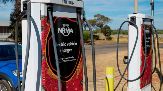 Two EV charging stations, decorated with Aboriginal art dot designs, along a sunny, rural road. 