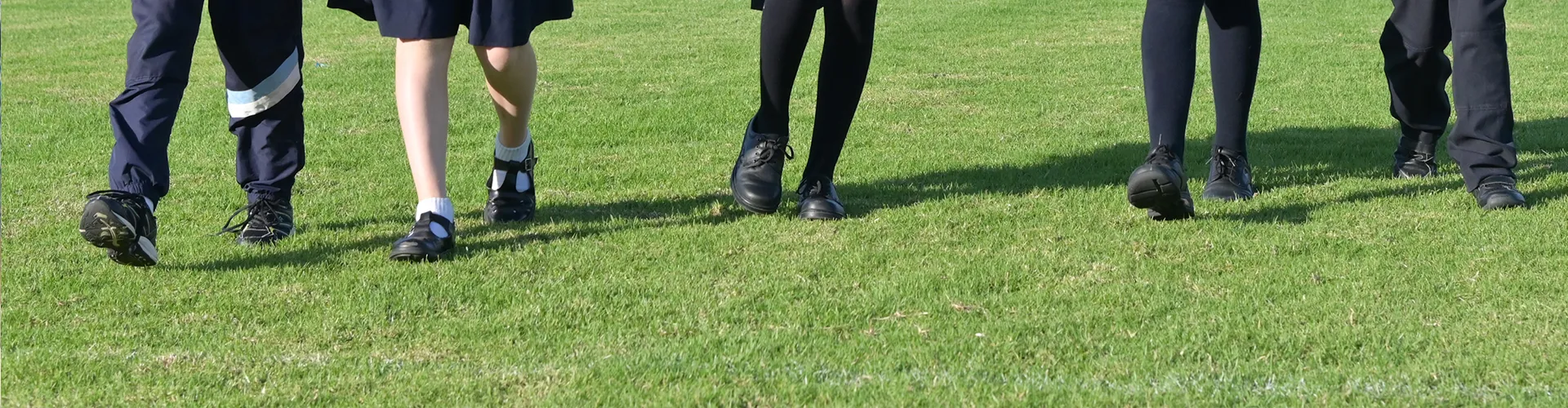 students walking across a grassy field while wearing school uniforms consisting of dark trousers, skirts, and black school shoes