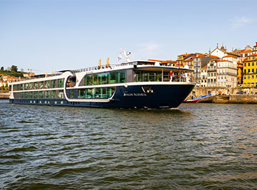 The Avalon Alegria river cruise ship sailing along a wide waterway, with large panoramic glass windows along its side.