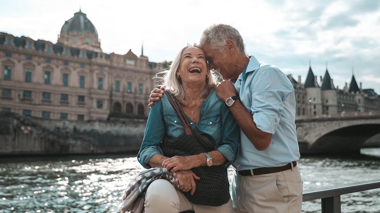 Elderly couple at a scenic riverfront with historic architecture and a landmark bridge.