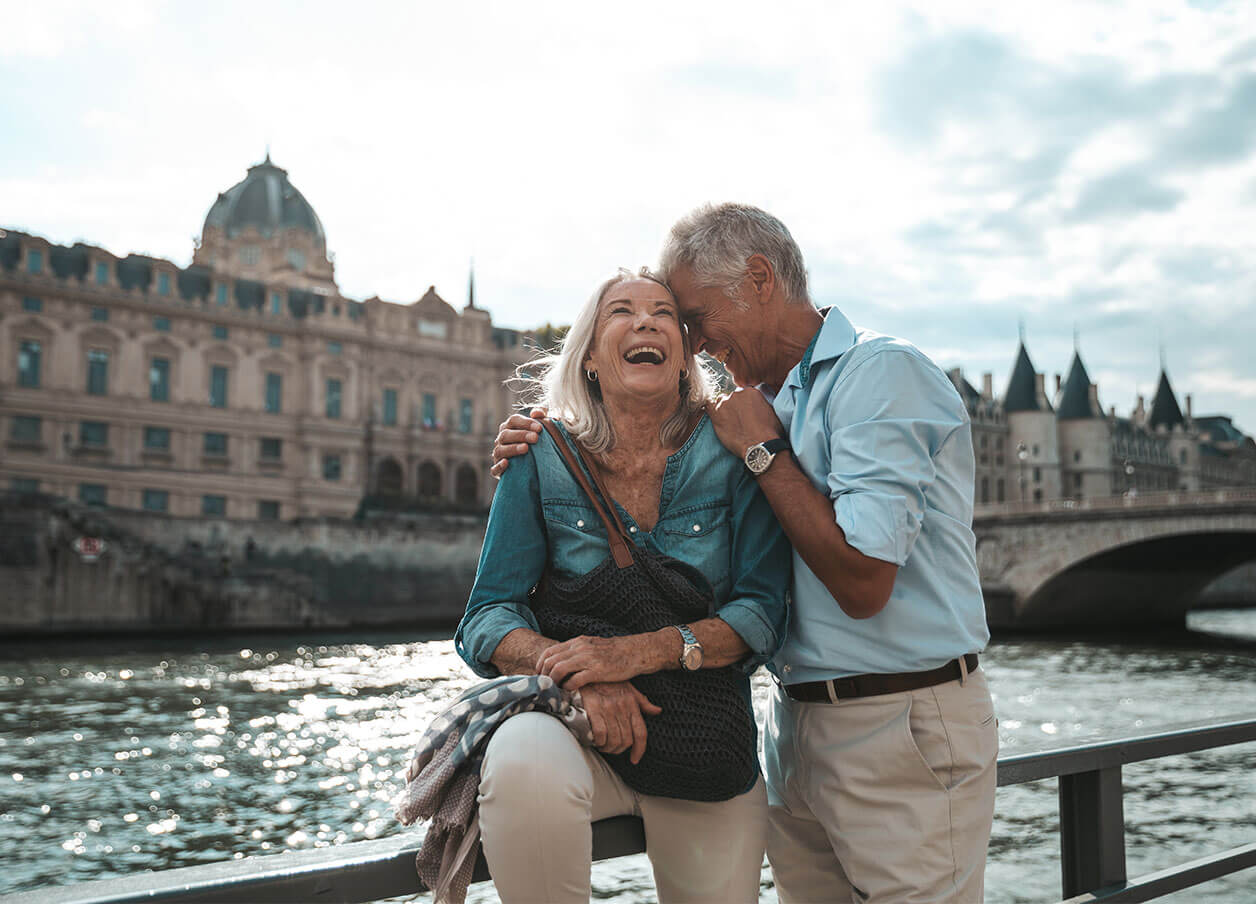 Elderly couple at a scenic riverfront with historic architecture and a landmark bridge.
