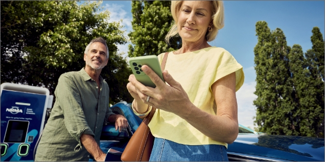 Woman in a yellow shirt looks at her phone while a man plugs an EV in