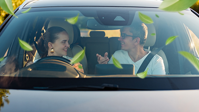 Two people smiling and chatting inside a car, seen through the windshield with green leaves drifting past.