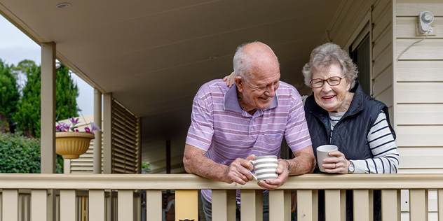 An old couple lean over the railing on their balcony with cups of tea