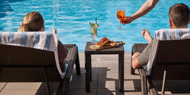 A man and woman on lounge chairs by the pool being served a drink
