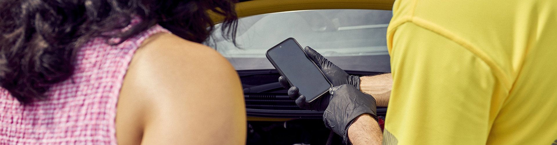 A woman and a man in an NRMA Roadside assistance uniform are examining a smartphone, showing curiosity and discussing what is displayed on a smartphone in relation to the vehicle in front of them