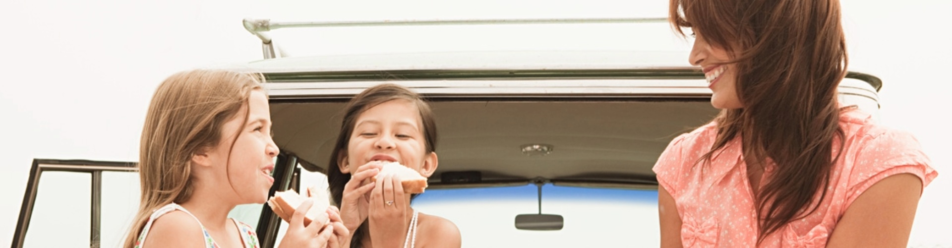 Eating healthy on a road trip woman and two young girls sit at the back of a vintage hatchback happily eating sandwiches