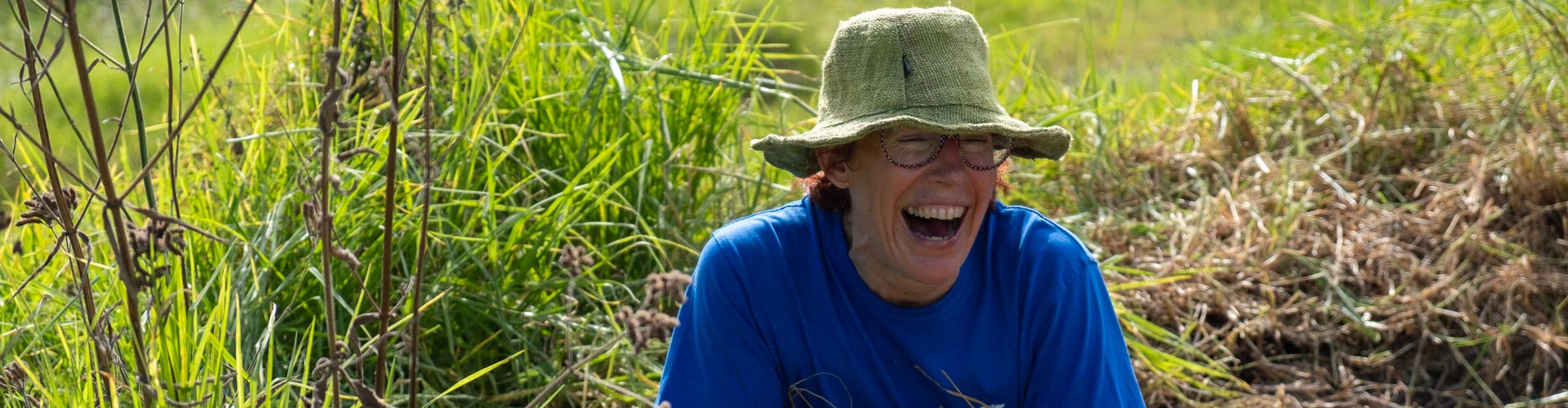 A woman wearing a blue t-shirt sits in the grass laughing on a sunny day