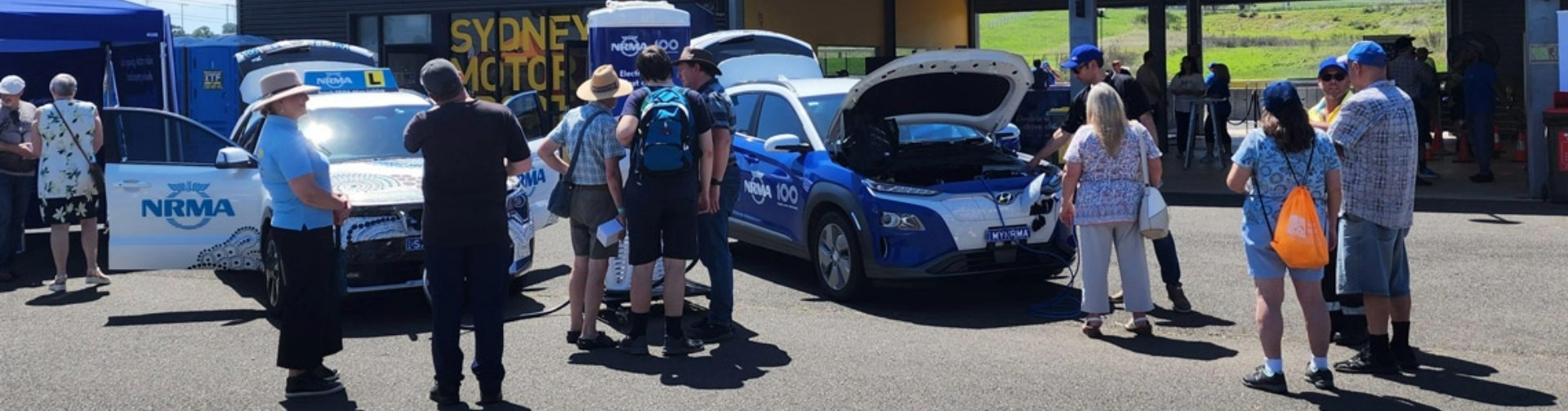 Members of the public at Sydney Motorsport Park complex, standing around two NRMA-branded electric vehicles and talking.