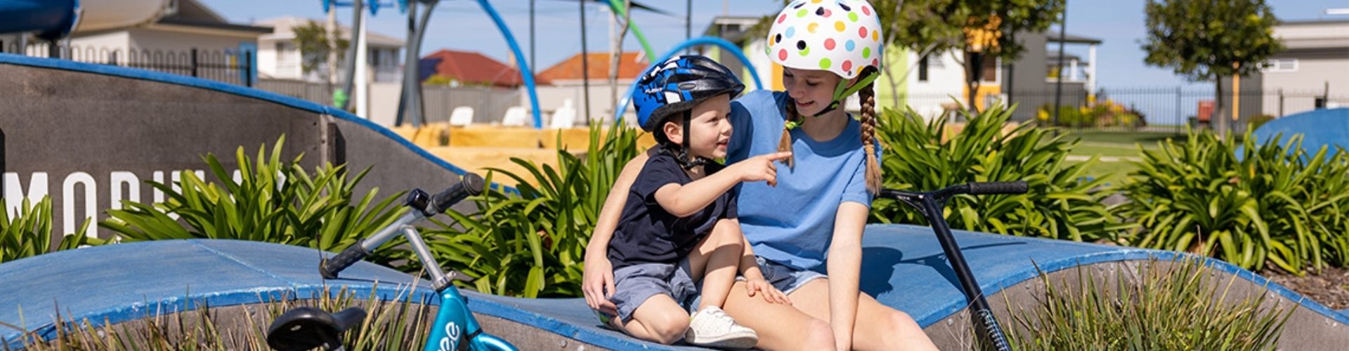 School holiday ideas - skate park small boy and teen girl with cycling helmets on sit on wave shaped skate ramp in play park