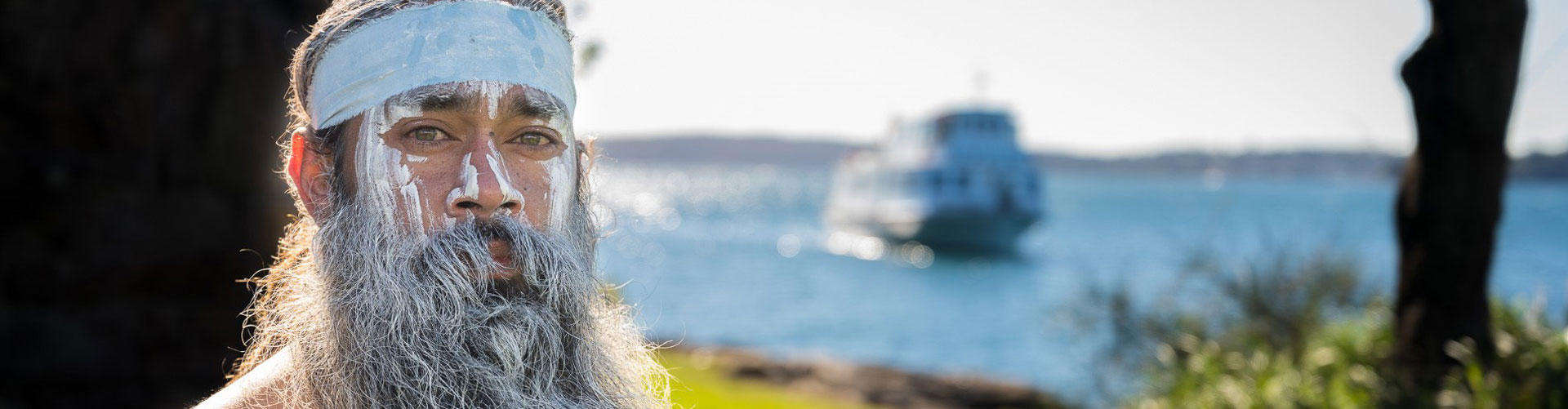 Australia Indigenous man staring at the camera with paint on his face standing in front of a blurry ferry and harbour in the background