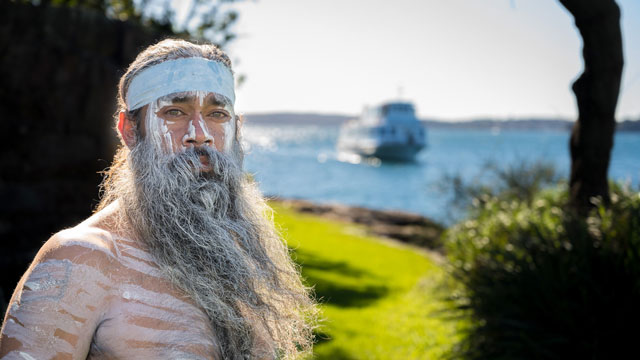 Australia Indigenous man staring at the camera with paint on his face standing in front of a blurry ferry and harbour in the background
