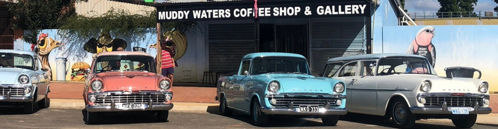 A row of vintage cars parked in front of a rustic-looking cafe with murals painted on the walls.