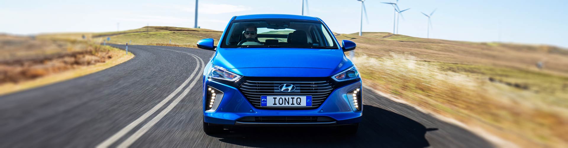 A blue Hyundai Ioniq hybrid driving towards the viewer with wind turbines behind