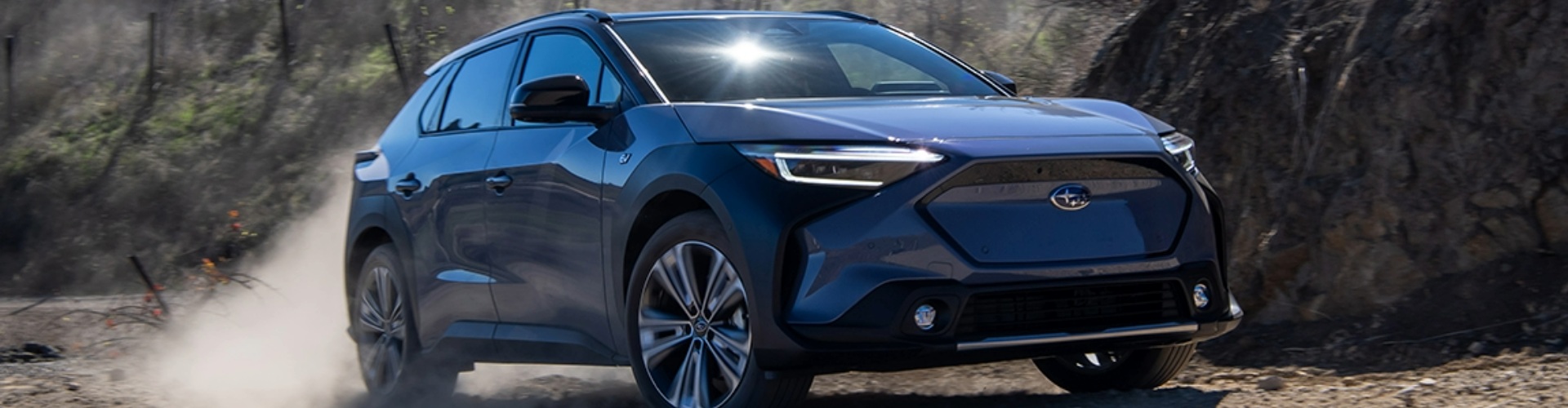 A blue 2023 Subaru Solterra EV driving along a dirt road with a rocky mountain backdrop.