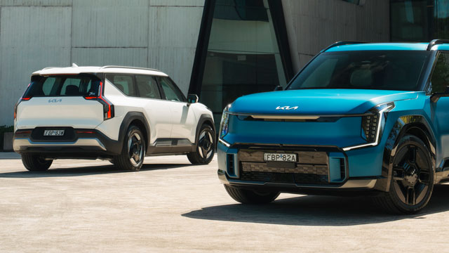A white, and a turquoise blue 2024 Kia EV9 SUV, parked in a sunny lot.