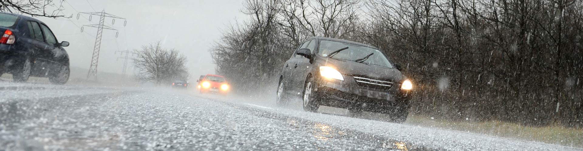 A black car driving in light hail on a wintry road with others behind it