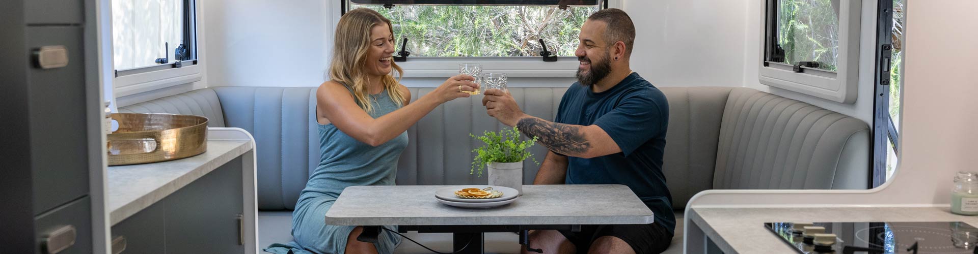 A couple clinks glasses behind a table in a caravan