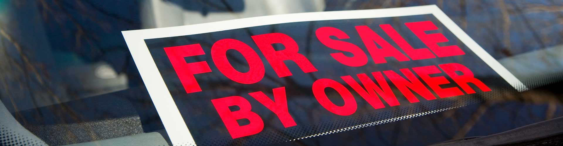A sign in car window reads For Sale by Owner in large red letters