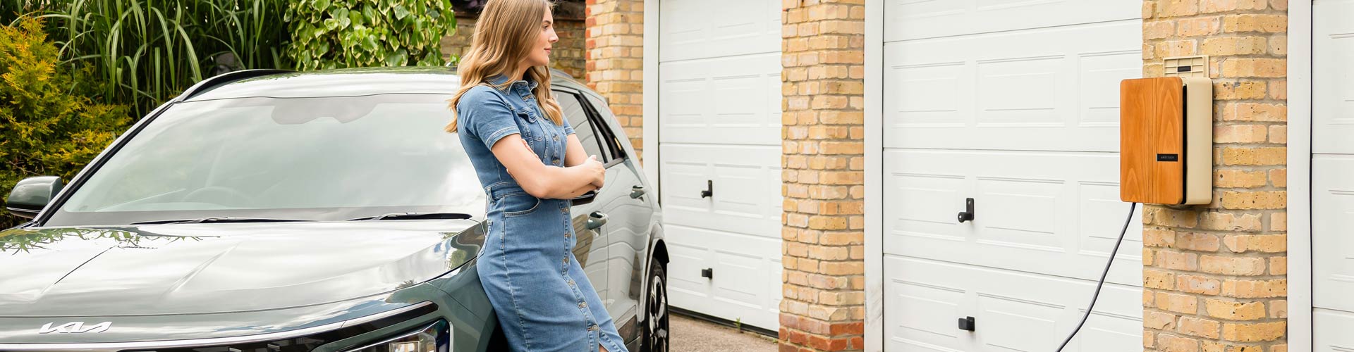 A woman in a denim dress leans on a blue Kia Niro EV while it charges next to a brick house