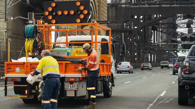 Workers stand near orange truck fixing road on Sydney Harbour Bridge