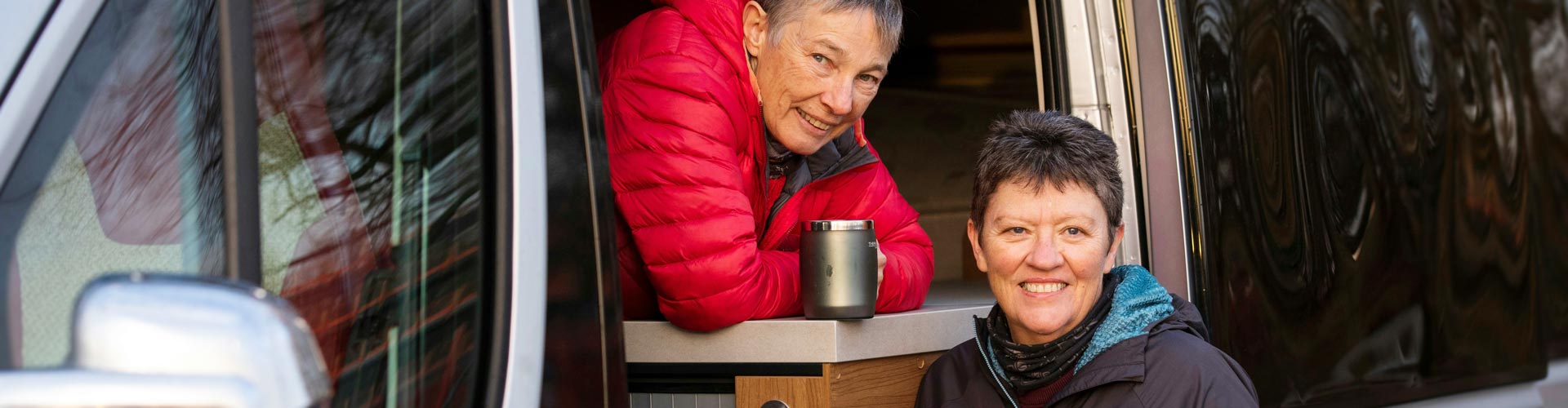 A woman holds a coffee cup in the window of a campervan while her partner stands outside the van