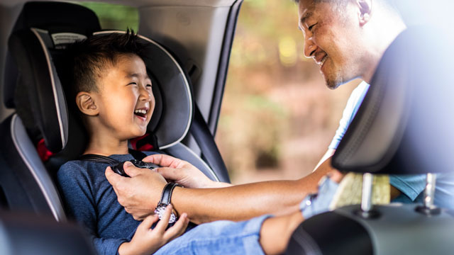 A father buckles his laughing child in to a car child seat in the back row