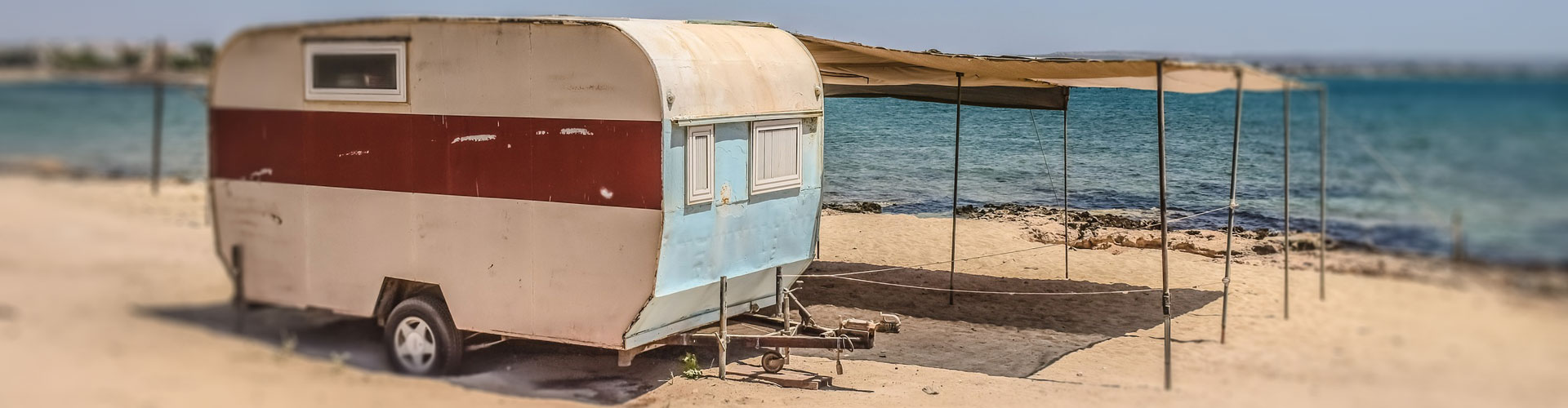 An old caravan with canvases attached by rope sits by a bay in front of the water
