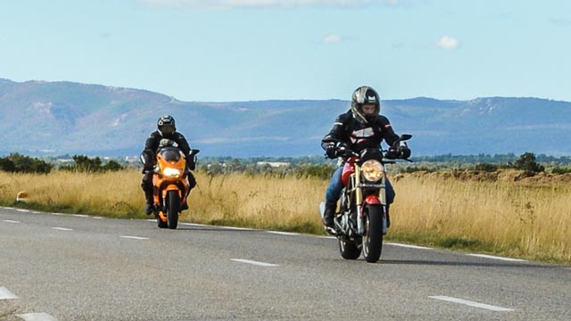 Two motorcyclist drive along a country road backed by mountains