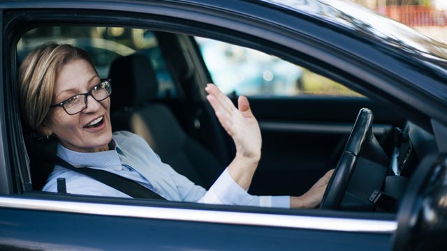 Middle-aged woman sitting in driving seat of car and waving to someone outside the car