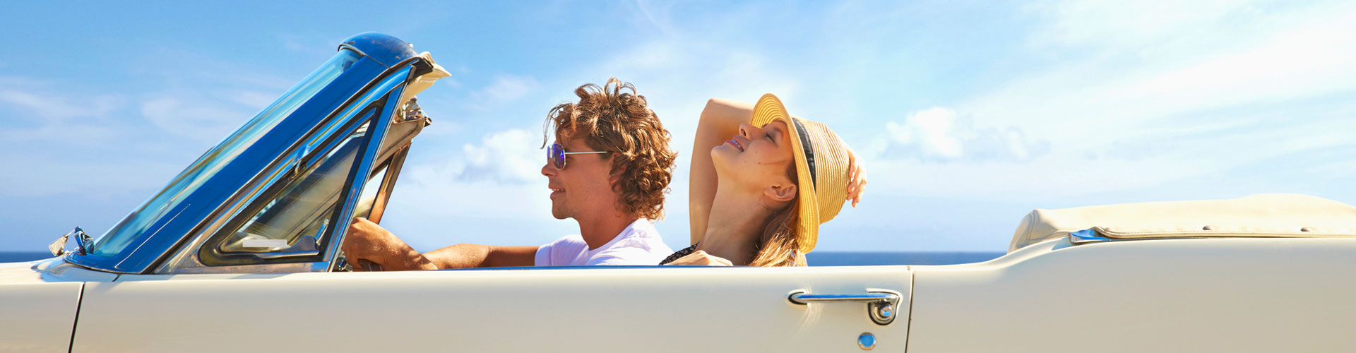 Young couple sitting in a cream convertible while the woman holds her hat on with her chin up