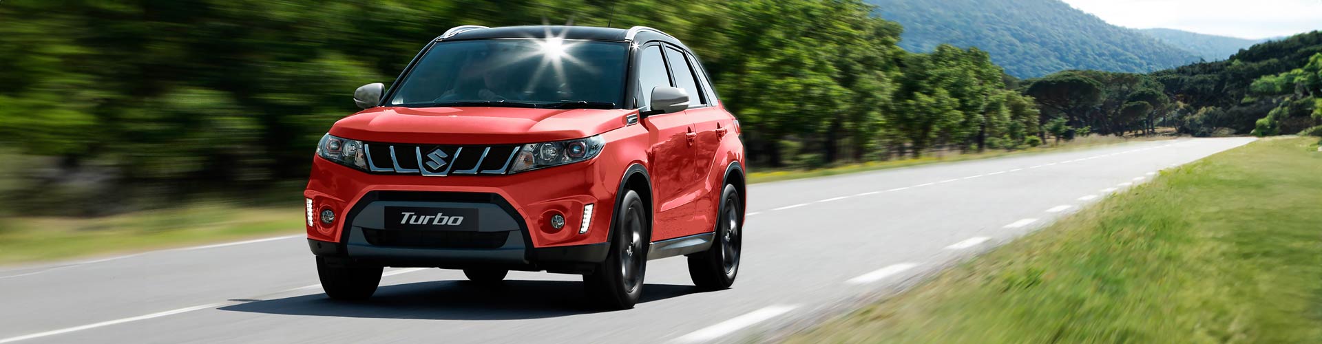 A red 2016 Suzuki Vitara S Turbo drives on a road edged by trees on a sunny day