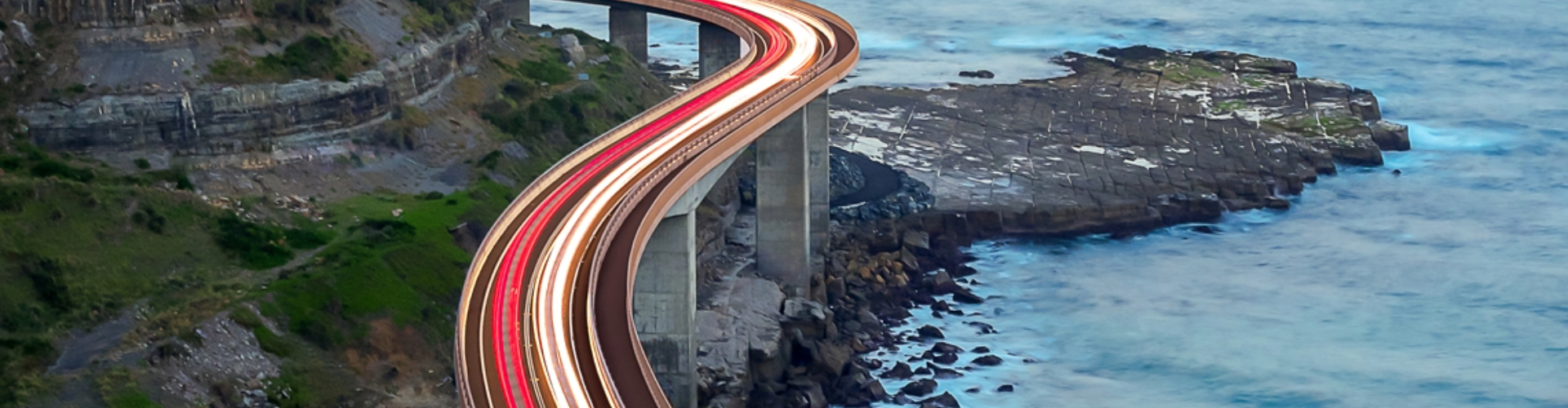 A trail of slow-exposure electric lights stream across an elevated ocean-side freeway