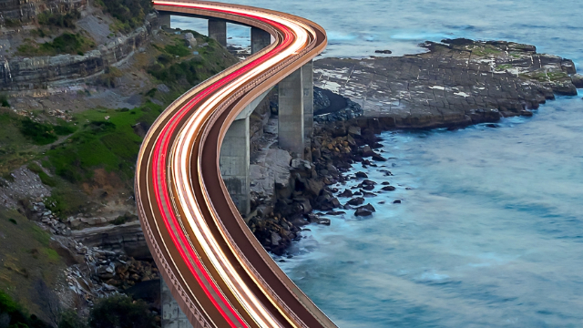 A trail of slow-exposure electric lights stream across an elevated ocean-side freeway