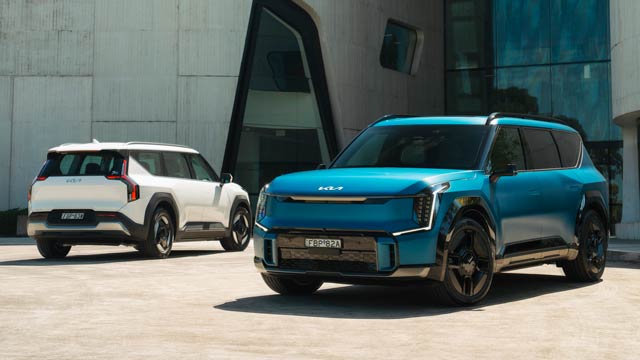 Two blue and white 2023 Kia EV9s parked in front of an angular building