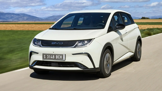 Front-angle view of a white BYD Dolphin driving along a road and a view of a picturesque background.