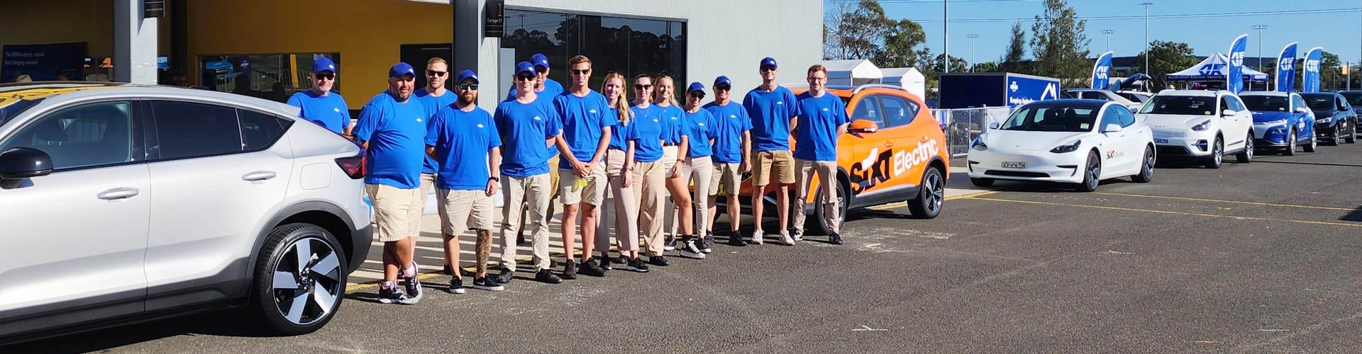 Row of staff members in blue NRMA shirts standing next to a line of parked electric cars